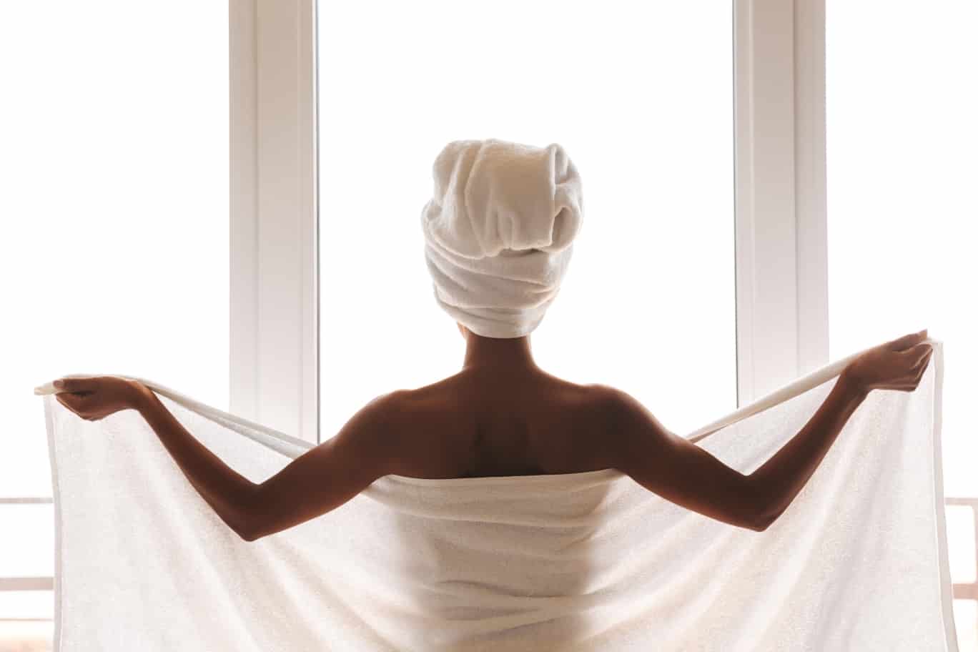 Frau steht mit geöffnetem weißen Saunahandtuch vor einer großen Fensterfront