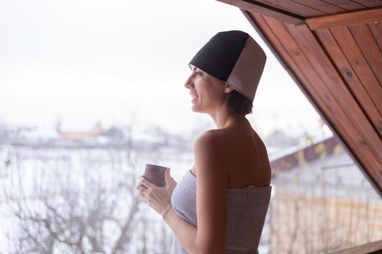 Young woman with a cap on her head, in a towel, holding tea, stands on the balcony in winter